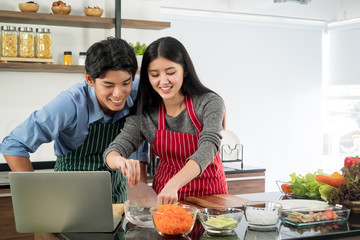 Happy and beautiful young couple cooking healthy food together, smiling while preparing vegetable salad meal in  kitchen and reading recipes on the laptop at home