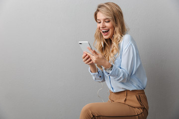 Canvas Print - Photo of alluring blond businesswoman with long curly hair smiling while listening to music with smartphone and earphones