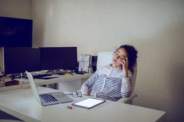 Tired businesswoman with curly brown hair sitting in chair at night and listening music over tablet. Office interior.