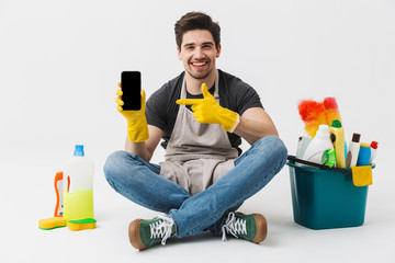 Wall Mural - Happy excited young houseman househusband with cleansers on floor isolated over white wall background showing display of mobile phone.