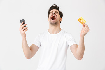 Wall Mural - Excited happy young man posing isolated over white wall background using mobile phone holding credit card.