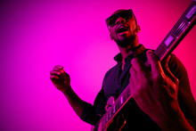 Young African-american Musician Playing The Guitar Like A Rockstar On Gradient Purple-pink Background In Neon Light. Concept Of Music, Hobby. Joyful Attractive Guy Improvising. Colorful Portrait.