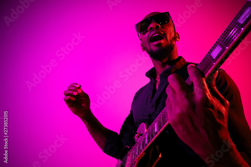 Obraz Dibond Młody afroamerykański muzyk grający na gitarze jak gwiazda rocka na gradientowym fioletowo-różowym tle w świetle neonowym. Pojęcie muzyki, hobby. Radosny atrakcyjny facet improwizujący. Kolorowy portret.
