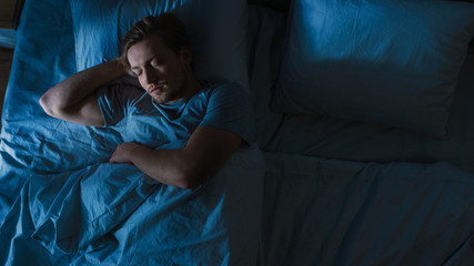 Top View of Handsome Young Man Sleeping Cozily on a Bed in His Bedroom at Night. Blue Nightly Colors with Cold Weak Lamppost Light Shining Through the Window.