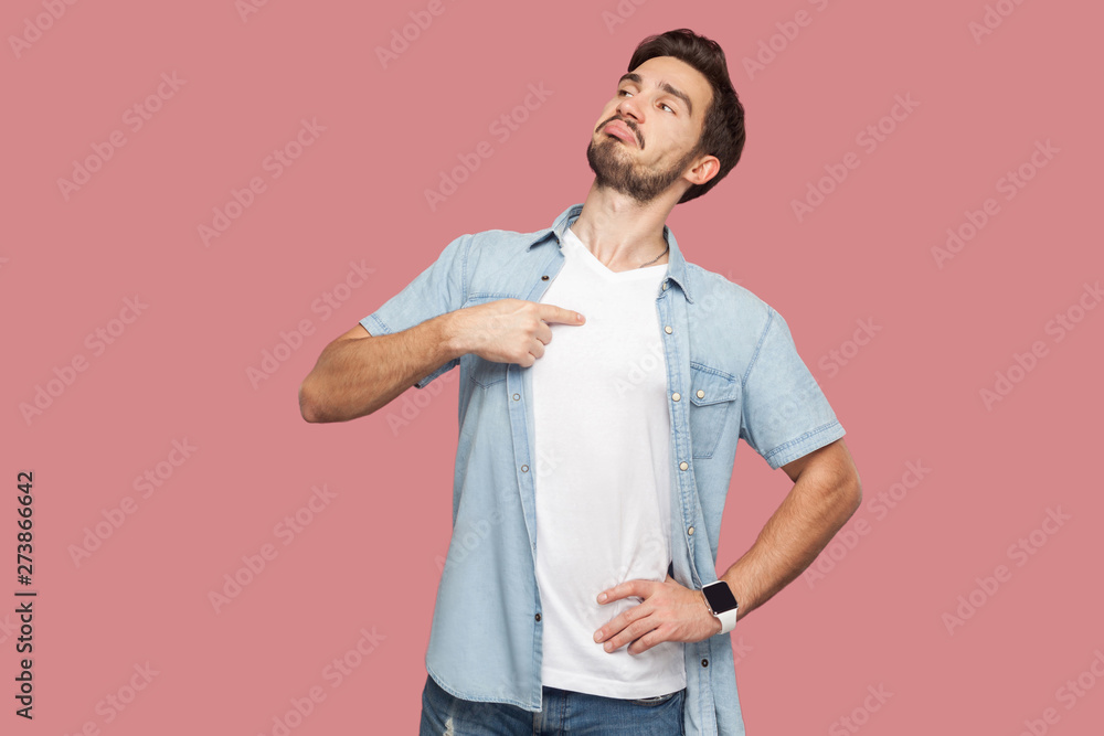 This is me. Portrait of proud haughty handsome bearded young man in blue casual style shirt standing, looking away and pointing himself. indoor studio shot, isolated on pink background. - obrazy, fototapety, plakaty 