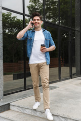 Poster - Image of joyful young man drinking takeaway coffee and talking on cellphone while standing over building