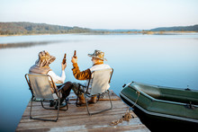 Grandfather With Adult Son Enjoying Beer, Sitting Together On The Pier While Fishing On The Lake Early In The Morning