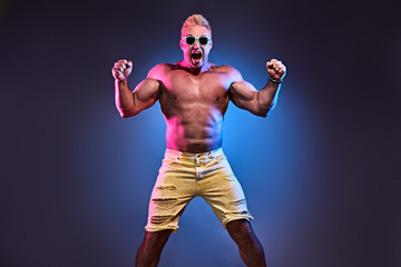 Excited Muscular strong expressive man with naked torso in fashion neon light. Sexy pumped-up bodybuilder fitness blonde guy in abs pose on blue. Male muscled torso. Strength and motivation. Creative