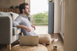 © Westend61 - Young man sitting at home on floor, using laptop, looking out of window