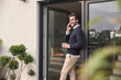 © Westend61 - Young man leaning in door of his house, holding cup of coffee, using smartphone