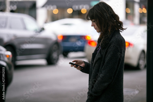Female pedestrian waiting for a rideshare.  She is sharing her gps location via cellphone app so the driver can pick her up in the city.  Cars are blurred to obscure make model and license plates.