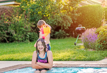 Happy siblings playing and havung fun in swimming pool. Little boy pouring water on his tween sister head from watering can. Summer lifestyle and leisure activity. Children enjoying summer holidays