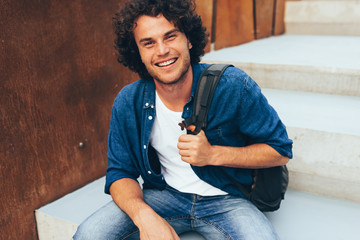 Portrait of happy smiling young man student ith backpack sitting at concrete stairs on the city street. Businessman freelancer male wearing casual outfit feelign positive. People, education, business.