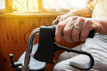 Aging woman holds her wrinkled hand on an adult medical walker sitting on the balcony in bright sunshine. Aging process and old age - old senior woman’s hand with wrinkled skin and veins.