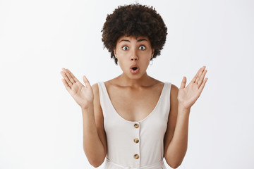 Wall Mural - Indoor shot of attractive african american female in stylish outfit with curly hairstyle raising palms and folding lips popping eyes from amazement and curiousity, being intrigued by fresh rumors