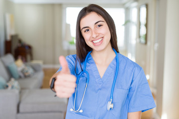 Sticker - Beautiful young nurse woman wearing uniform and stethoscope at the clinic doing happy thumbs up gesture with hand. Approving expression looking at the camera with showing success.