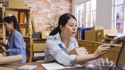 Asian businesswoman hold small cardboard box. Online shopping young start small business checking order with parcel at work on laptop. two seller prepares delivery packages for customer online sales.