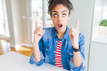 Beautiful young african american woman with afro hair wearing casual denim jacket amazed and surprised looking up and pointing with fingers and raised arms.