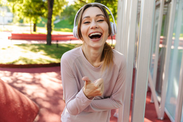 Poster - Beautiful cheerful young fitness sports woman posing outdoors in park listening music with earphones.