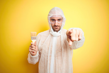 Sticker - Young man wearing painter equipment and holding painting brush over isolated yellow background pointing with finger to the camera and to you, hand sign, positive and confident gesture from the front