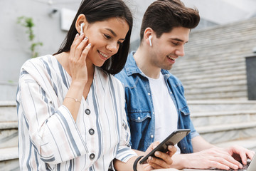 Wall Mural - Happy young amazing loving couple business people colleagues outdoors outside on steps using mobile phone and laptop computer listening music with earphones.