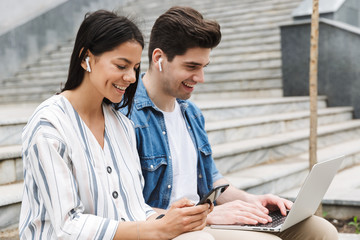 Canvas Print - Happy young amazing loving couple business people colleagues outdoors outside on steps using mobile phone and laptop computer listening music with earphones.