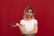 © ianachyrva - Portrait of a girl with curly blond hair in a white t-shirt on a red background. Unhappy model in wireless headset holds mobile phone.