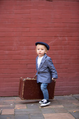 Poster - Cute child, boy in vintage cloths, eating lollipop ice cream, sitting on vintage suitcase in front of a red brick wall