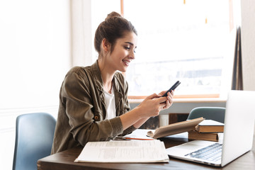 Canvas Print - Beautiful happy young concentrated woman using laptop computer indoors using mobile phone chatting.