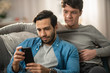 © Gorodenkoff - Cute Male Gay Couple Spend Time at Home. They are Lying Down on a Sofa and Use a Smartphone. They Browse Online. Partner's Hand is Around His Lover. They Smile and Laugh. Room Has Modern Interior.