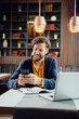 © chika_milan - Young Caucasian businessman dressed smart casual using smart phone while sitting in cafe. On the desk is laptop.