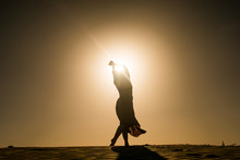 Silhouette Of Young Woman With Long Skirt Dancing In Evocative And Confident Way On Top Of Desert Dune At Sunset With Sun High In The Sky