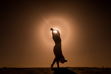 Silhouette Of Young Woman With Long Skirt Dancing In Evocative And Magic Way On Top Of Desert Dune At Sunset With Sun High In The Sky