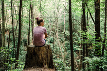 Teen Teen Girl Sitting On A Stump Looking Out Into A Green Forest Full Of Trees