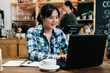 © PR Image Factory - Image of happy asian young woman using laptop computer while sitting at cafe bar. japanese girl sitting in coffee shop and working on notebook pc. female barista in counter making cooking indoors.