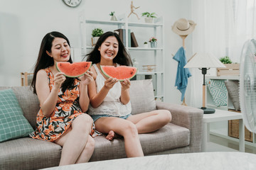 leisure and friendship concept. happy smiling teenage girls friends eating watermelon in summer house indoor. young asian women sitting on couch sofa enjoy fresh fruits at home laughing hot outdoor