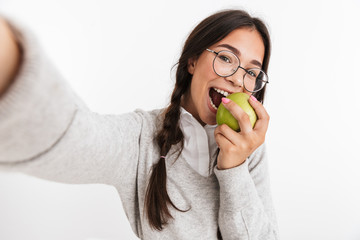 Poster - Happy young school girl isolated over white wall background posing take a selfie by camera eat apple.