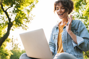 Wall Mural - Image of pleased caucasian woman using silver laptop while sitting on bench in green park