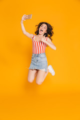 Sticker - Portrait of excited brunette woman in denim skirt jumping and showing thumb up while taking selfie on cellphone