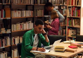 Wall Mural - Young male student study in the library using laptop for researching online, while his friend searching the book on the bookshelf behind him.