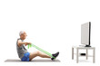 © Ljupco Smokovski - Elderly man sitting and exercising with an elastic band in front of a tv