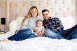© Cavan Images - Mom dad and baby sitting on bed and smiling at camera