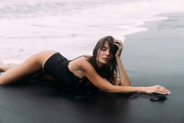 Wall Mural - Sexy sensual girl with wet hair in swimwear lies on deserted black sandy beach