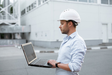Young engineer in hardhat standing on the background of water or gas pipes station and using laptop. Heating station manager doing his job outdoor.