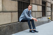 © Alexander Image - Young European Businessman traveling in New York City, wearing gray blazer, white shirt, black tie, pants, leather shoes, sitting on old street on window frame against wall, relaxing, waiting..