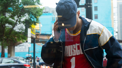 Chicago, IL/United States-June 12th 2019:  Young attractive Middle eastern male model is talking on his smart phone smiling while wearing fashion trendy clothes. view from a inside a window pane