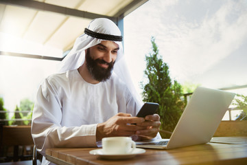 Arab saudi businessman working online with a laptop and tablet in a coffee shop or a cafe with an outdoor terrace in the background. Concept of business, finance, modern technologies, start up.