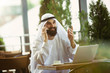 © master1305 - Arab saudi businessman working online with a laptop and tablet in a coffee shop or a cafe with an outdoor terrace in the background. Concept of business, finance, modern technologies, start up.