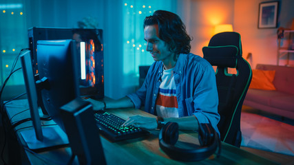 Wall Mural - Excited Gamer Playing Online Video Game on His Personal Computer. Room and Personal Computer have Colorful Warm Neon Led Lights. Young Man has Long Hair and Handsome Smile. Cozy Evening at Home.