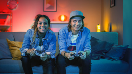 Wall Mural - Excited Black Gamer Girl and Young Man Sitting on a Couch and Playing Video Games on Console. They Plays with Wireless Controllers. Cozy Room is Lit with Warm and Neon Light.